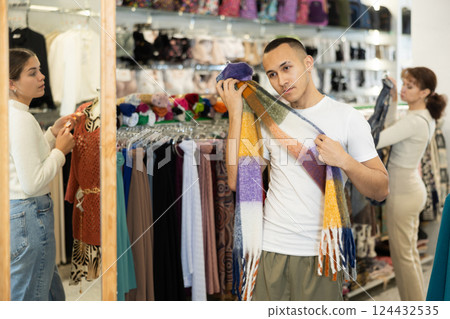 Guy stopped near accessories showcase, holds scarf hanger in hands, examines product 124432535