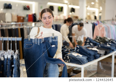 Girl buyer in store has removed hanger with jeans from window and is considering product. 124432645