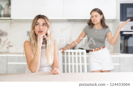 Middle-aged woman sitting at the table while a girl is disputing with her standing behind Middle-aged woman sitting at the table while a girl is disputing with her standing behind 124433004
