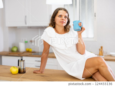 Portrait of young woman in white dress drinking morning coffee in modern kitchen Portrait of young woman in white dress drinking morning coffee in modern kitchen 124433136