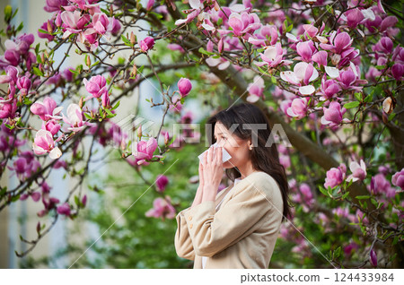Woman allergic suffering from seasonal allergy at spring in blossoming garden at springtime. Young woman sneezing and blowing nose with nasal handkerchief in front of blooming tree. Allergy concept. Woman allergic suffering from seasonal allergy at spring in blossoming garden at springtime. Young woman sneezing and blowing nose with nasal handkerchief in front of blooming tree. Allergy concept. 124433984