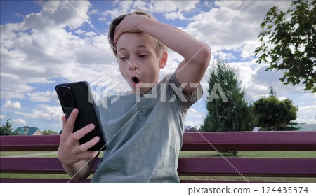 One teenager young man caucasian guy is sitting on a bench in the park and using his phone. The emotions on the face are Surprise and delight from the message on the phone. Teen boy uses phone One teenager young man caucasian guy is sitting on a bench in the park and using his phone. The emotions on the face are Surprise and delight from the message on the phone. Teen boy uses phone 124435374