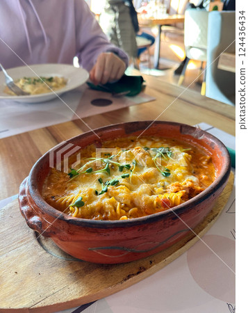 Baked pasta served in clay dish on wooden board. Conveys warmth, tradition, and the comfort of a hearty homemade-style meal in a cozy setting. 124436434