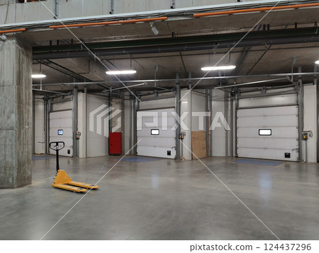 Spacious warehouse loading dock with closed roll-up doors, industrial lighting, and a yellow pallet jack on a polished concrete floor. 124437296