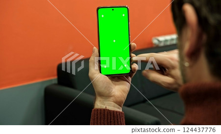 Hand of young man holding smartphone green screen in business office work. Mobile phone vertical mockup. Male person using phone with blank green background display. Chroma key smart phone. Close up. 124437776
