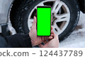 Male hands holding a smartphone with a bright green screen against the background of a car wheel. Automotive technology. Mobile applications for drivers. Road safety. Using gadgets. Chroma key 124437789