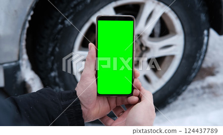 Male hands holding a smartphone with a bright green screen against the background of a car wheel. Automotive technology. Mobile applications for drivers. Road safety. Using gadgets. Chroma key 124437789