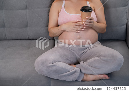 Faceless pregnant woman in 3rd trimester holding a coffee glass on her belly.  124437823