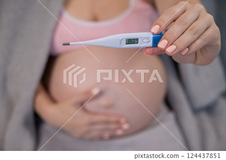 Pregnant woman measures temperature while sitting on the couch. Close up of the belly. 124437851