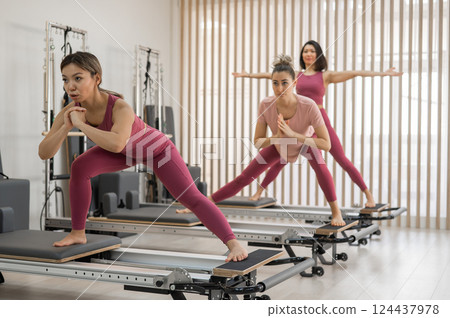 Three Asian women in pink sportswear doing pilates exercises with a reformer bed. Three Asian women in pink sportswear doing pilates exercises with a reformer bed. 124437978