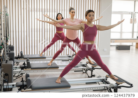 Three Asian women in pink sportswear doing pilates exercises with a reformer bed. Three Asian women in pink sportswear doing pilates exercises with a reformer bed. 124437979