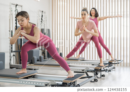 Three Asian women in pink sportswear doing pilates exercises with a reformer bed.  124438155