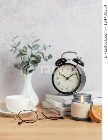 Alarm clock, books, coffee cup, eyeglasses and candle creating cozy atmosphere on wooden desk 124438214