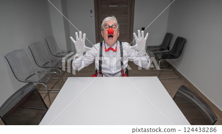 An elderly man in a clown costume sits at a desk in an office.  124438286