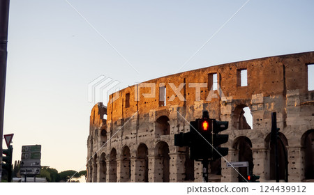 A view of the Colosseum in Rome bathed in warm sunset light, with a traffic light in the foreground, blending history and modernity. 124439912