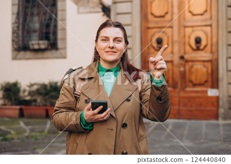 Happy cheerful young woman with backpack walking on city street checks her smartphone. Portrait of beautiful 30s girl using smartphone outdoors. Urban lifestyle concept. 124440840