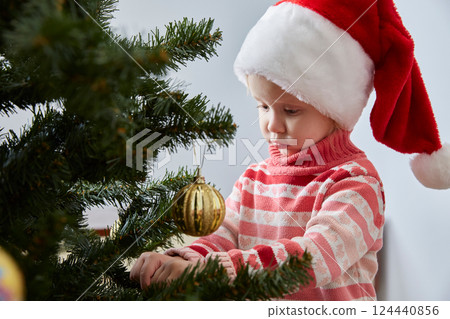 girl in a Santa hat hangs a Christmas ball on a Christmas tree girl in a Santa hat hangs a Christmas ball on a Christmas tree 124440856