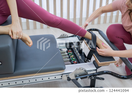 Personal Pilates training. Instructor adjusts woman on reformer.  124441094