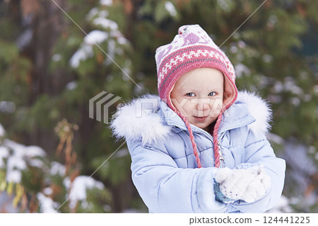 A smiling girl in a pink hat and a blue jacket with fur. In winter, he holds snow in his hands. Looks at the camera. Place for text, banner, new year. 124441225