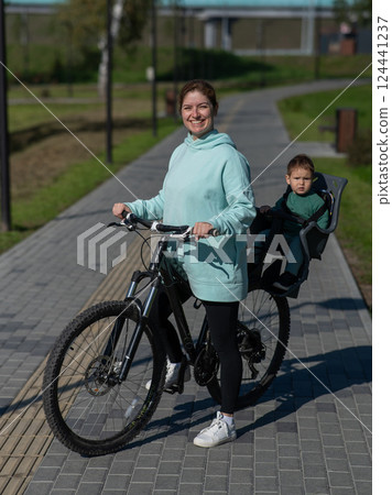 Caucasian woman riding bicycle with toddler son sitting behind her in child seat in park. Vertical photo.  124441237