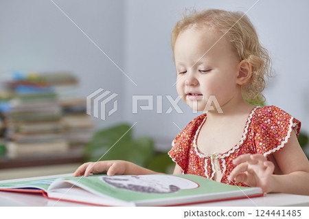 Curly-haired cute girl toddler reading book. Close-up. Room looks like house or library. 124441485