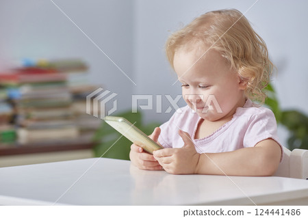 child girl is sitting at a table with a phone in her hands and smiling while looking at a smartphone. 124441486