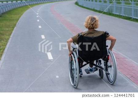 Rear view of an elderly woman in a wheelchair riding on a bike path. Rear view of an elderly woman in a wheelchair riding on a bike path. 124441701