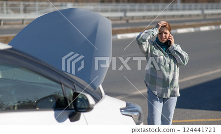 Caucasian woman cries and calls on the phone standing near the car with the hood open. Caucasian woman cries and calls on the phone standing near the car with the hood open. 124441721