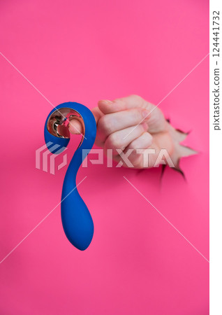 Hand with blue vibrator sticking out through pink paper background. Vertical photo. Hand with blue vibrator sticking out through pink paper background. Vertical photo. 124441732