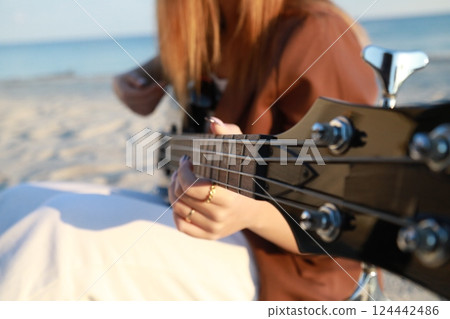 Girl Plays The Bass Instrument Sitting On The Beach At Sunset Near The Ocean 124442486