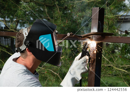 Man welds iron fence outdoors. Backside view. Welder repairs makes iron fence. Sunny day, green hedge background. Construction. Summer. Outdoors. 124442556
