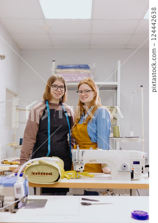 Two female seamstresses in workshop at factory are standing at a table with sewing machines.  Two female seamstresses in workshop at factory are standing at a table with sewing machines.  124442638