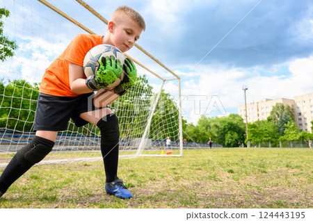 Boy in orange shirt and green gloves holding football with both hands, standing in front of goal. Boy in orange shirt and green gloves holding football with both hands, standing in front of goal. 124443195