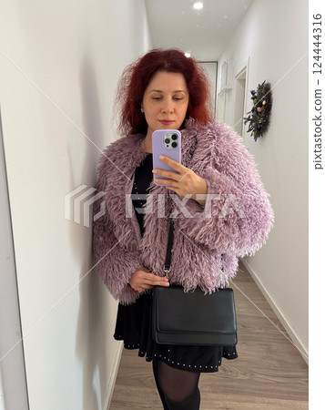 Woman in textured fur coat taking mirror selfie in hallway. Self-expression, fashion, and everyday life in a home environment. 124444316