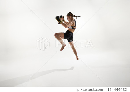 Focused athlete in black boxing gear performing high jump for attack move, training against white background. 124444482
