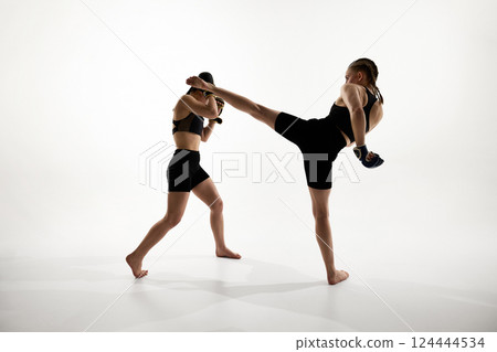 Strong female athletes in active stance, executing high kick and blocking punch during martial arts training against white background 124444534