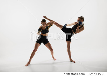 Two athletic female fighters in black sportswear and boxing gloves training powerful high kick move during sparring against white background 124444536