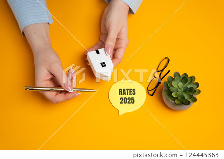 Female hands is a miniature house near notes with the words Rates 2025. Loan and credit interest. Apartments, housing, house. Real estate market analysis concept. Business and Finance 124445363