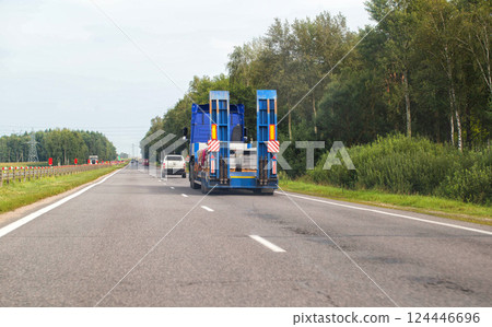 A blue truck tractor with a semi-trailer transports cargo along a country highway against the backdrop of a forest in the summer. Copy space for text, freight transport company 124446696