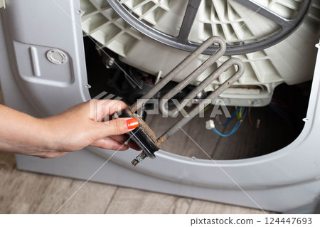 A woman's hand holds an electric heating element for heating water against the background of a washing machine. Heating element breakdown concept, close-up A woman's hand holds an electric heating element for heating water against the background of a washing machine. Heating element breakdown concept, close-up 124447693