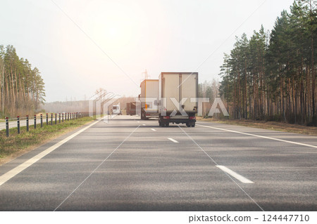 Traffic from trucks transporting goods along a country asphalt road against the backdrop of forest and sun in spring. Economics and business of cargo transportation. Copy space for text Traffic from trucks transporting goods along a country asphalt road against the backdrop of forest and sun in spring. Economics and business of cargo transportation. Copy space for text 124447710