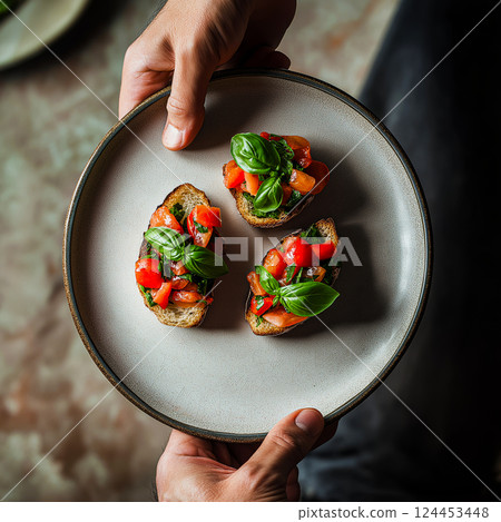 Chef holding plate with two slices of toasted bread topped with fresh tomatoes, basil, and seasonings, creating a classic italian bruschetta appetizer 124453448