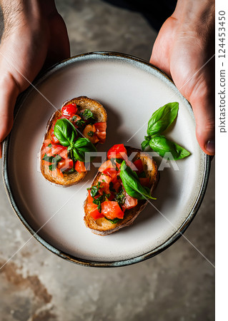 Chef holding plate with two slices of toasted bread topped with fresh tomatoes, basil, and seasonings, creating a classic italian bruschetta appetizer 124453450