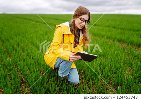 Young farmer woman in a green field, intently using a tablet. She wears a yellow jacket and glasses Young farmer woman in a green field, intently using a tablet. She wears a yellow jacket and glasses 124453748