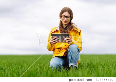 Young farmer woman in a green field, intently using a tablet. She wears a yellow jacket and glasses 124453749