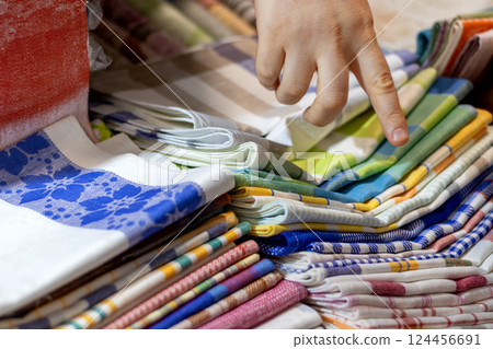 Shopper's hand points to selected item in colorful stack of folded fabrics on market stall. Variety of textile options for kitchen napkins and towels 124456691