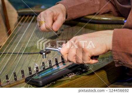Close-up of man's hands tuning traditional stringed instrument using tuning tool and smartphone app. Psaltery Close-up of man's hands tuning traditional stringed instrument using tuning tool and smartphone app. Psaltery 124456692