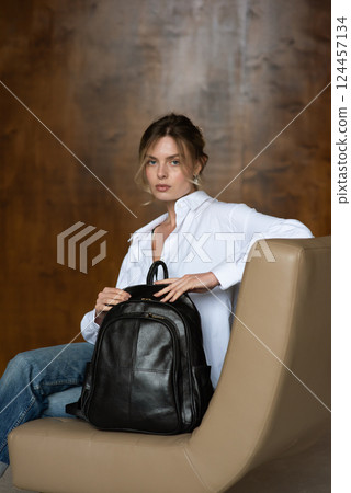 A girl in a white shirt and blue jeans is posing with a black backpack. 124457134