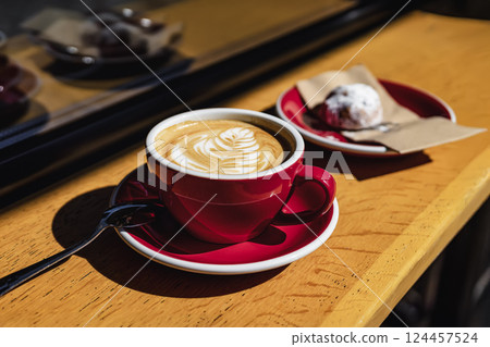 Red coffee cup with creamy latte art sits in golden sunlight beside cookie on napkin. Coffee moment feels warm and inviting with cozy atmosphere and perfect coffee pairing 124457524