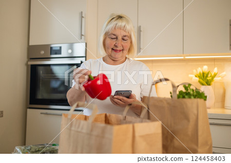 Senior woman holding red bell pepper while checking phone in the kitchen Senior woman holding red bell pepper while checking phone in the kitchen 124458403